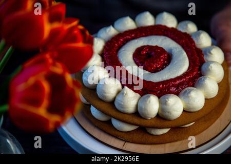 Der gebackene Kuchen liegt auf einem Holzsubstrat und auf einem weißen Stand, der Kuchen ist mit weißer Creme und Himbeermarmelade dekoriert, in einem Kreis, abwechselnd Kreise, ein Strauß roter Tulpen auf dunklem Hintergrund ist in der Nähe sichtbar Stockfoto