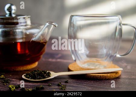 Ein Holzlöffel mit Tee liegt vor einem dunklen Holzhintergrund, ein Becher und eine Teekanne sind von hinten sichtbar, ein Foto in dunklen Farben. Stockfoto