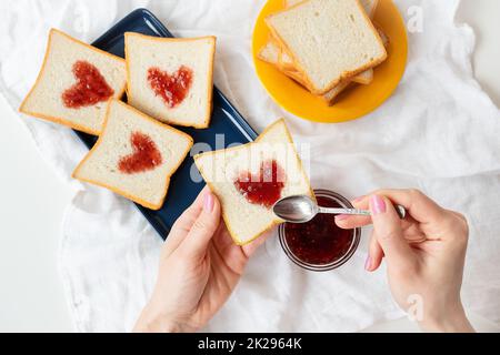Das Mädchen macht einen Toast, auf dem das Herz aus Marmelade besteht. Überraschungsfrühstück im Bett. Romantik zum Valentinstag Stockfoto