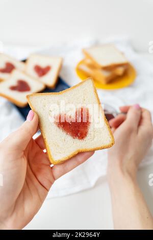 Das Mädchen macht einen Toast, auf dem das Herz aus Marmelade besteht. Überraschungsfrühstück im Bett. Romantik zum Valentinstag Stockfoto