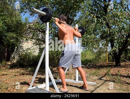 Der Mann nimmt eine Bar, um Squats zu machen. Übungen im Bodybuilding. Sport im Garten. Stockfoto