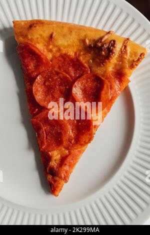 Eine Scheibe Pepperoni-Pizza auf weißem Keramikplatte, die wie ein Pappteller aussieht Stockfoto