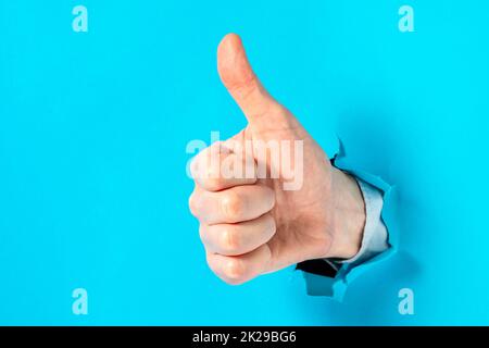 Nahaufnahme der Hand eines Mannes mit Daumen-hoch-Geste durch blaues Loch in der Papierwand Stockfoto