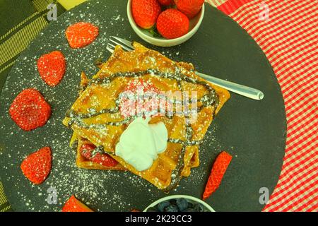 Traditionelle belgische Waffeln mit Heidelbeeren und Erdbeeren. Geschmolzene Schokolade und Schlagsahne auf Waffeln. Draufsicht. Schwarzer Schieferhintergrund Stockfoto