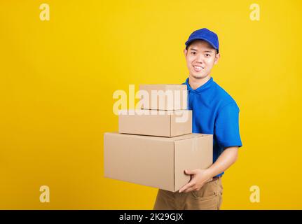 Portrait aufgeregt attraktive Lieferung glücklich Mann logistischen stehend er lächeln in blauen T-Shirt und Mütze Uniform halten Paketkasten Blick auf die Kamera, Studioaufnahme isoliert auf gelbem Hintergrund Stockfoto