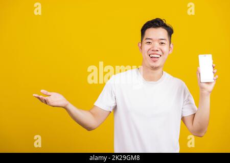 Ein gut aussehender, asiatischer junger Mann, der positiv lächelt, zeigt einen leeren Bildschirm mit einer anderen Handfläche, einen aufgeregten, glücklichen männlichen Hold Smartphone Studio, isoliert auf gelbem Hintergrund aufgenommen Stockfoto