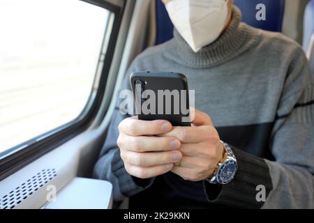 Mann auf öffentlichen Verkehrsmitteln mit mobiler App trägt medizinische Gesichtsmaske. Pendler mit Mobiltelefon und obligatorischer Schutzmaske KN95 FFP2. Konzentrieren Sie sich auf das Telefon. Stockfoto