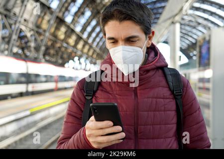 Junger Reisender Mann mit KN95 FFP2 Gesichtsmaske am Bahnhof. Junger kaukasischer Mann, der im Bahnhof ein Smartphone benutzt. Stockfoto