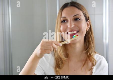 Eine junge Frau mit einer Zahnbürste aus Holzbambus im Bad, die sich selbst in den Spiegel schaut. Zahnpflege, umweltfreundliche Bambuszahnbürste, keine Verschwendung. Stockfoto