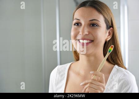 Eine junge lächelnde Frau hält eine Bambuszahnbürste und sieht sich zu Hause in den Spiegel. Zahnpflege, umweltfreundliche Bambuszahnbürste, keine Verschwendung. Stockfoto