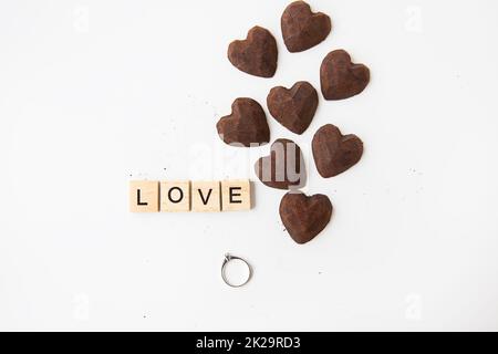 Die Inschrift in hölzernen Buchstaben liebt kleine Trüffelbonbons in Form eines Herzens. Ring mit Diamant - ein Vorschlag von Hand und Herz. Stockfoto