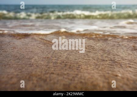 Sehr niedriger Winkel (Kamera auf dem Boden, Nahaufnahme) Meerwasser wäscht den Sand. Selektiver Fokus auf den nassen Strand, abstrakter Sommerurlaub / Urlaubshintergrund. Stockfoto