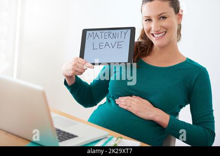 Ich freue mich darauf, Mutter zu sein. Eine Schwangerin, die eine Tablette mit den Worten Mutterschaftsurlaub auf dem Bildschirm hält. Stockfoto