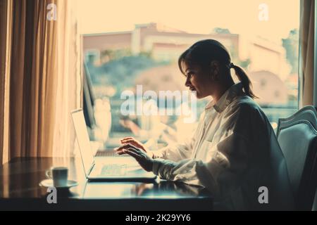 Seitenansicht einer schönen erwachsenen Latinerin, die in einem Coworking Space neben dem Fenster sitzt und ihren Laptop benutzt; ein Porträt einer hispanischen Frau, die es benutzt Stockfoto