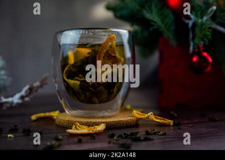 Eine durchsichtige Tasse Tee steht auf einem dunklen Tisch, auf einem Holzständer, getrocknete Orangenscheiben schwimmen in der Tasse, Zitrusscheiben liegen daneben, Tee ist verstreut, ein Foto in einem dunklen Schlüssel Stockfoto