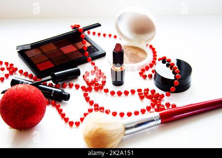 Puder und Bronzer, Lidschatten, Lippenstift, Rouge und Pinsel verschiedener Marken liegen auf weißem Hintergrund, umgeben von roten Weihnachtsbällen. Stockfoto