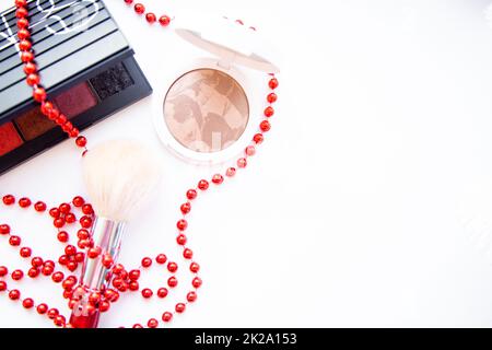 Eine weiße Pillbox mit Pulver und Bronzer, ein schwarzes Paket mit Augenschatten und einem Pinsel liegen auf weißem Hintergrund und sind mit roten Perlen verflochten, mit einem Platz für Text Stockfoto