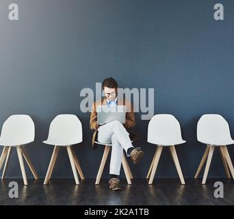 Das Warten lohnt sich. Studioaufnahme eines jungen Geschäftsmannes, der einen Laptop benutzt, während er vor grauem Hintergrund in der Schlange wartet. Stockfoto