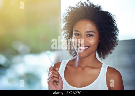 Arbeiten für dieses gewinnende Lächeln. Beschnittenes Porträt einer jungen Frau, die sich im Badezimmer die Zähne putzt. Stockfoto