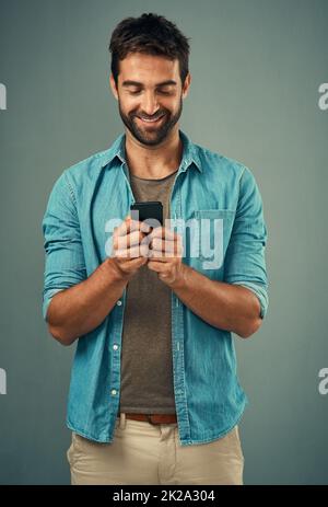 Hes ist derjenige, mit dem jeder in Kontakt sein möchte. Studioaufnahme eines hübschen jungen Mannes, der ein Mobiltelefon vor grauem Hintergrund verwendet. Stockfoto