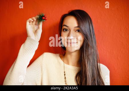 Bedeutet das, dass ich einen Kuss bekomme. Eine attraktive junge Frau, die einen Stechenzweig hochhält, während sie gegen eine rote Wand steht. Stockfoto