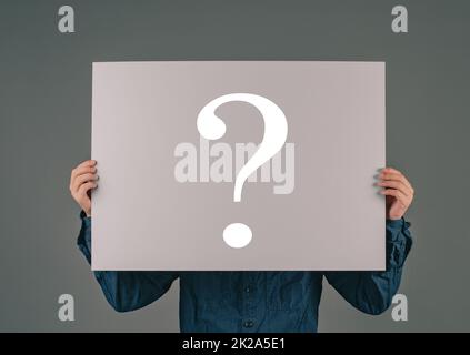 Ein junger Mann hält ein großes Papier mit einem Fragezeichen, fragt warum, sucht nach Antworten, dunkler Hintergrund Stockfoto