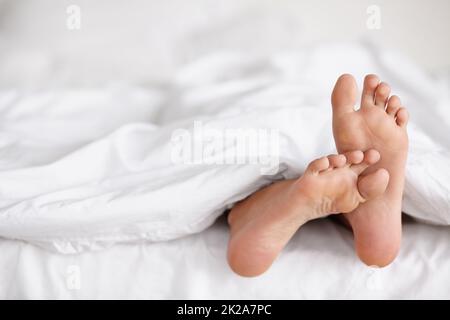 Wackelnde Zehen der Wachsamkeit. Aufnahme eines weiblichen Fußes, das unter der Bettwäsche eines Bettes herausstoss. Stockfoto