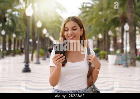 Mittelstarke junge lächelnde Frau nutzt Smartphone liest Nachrichten aus sozialen Netzwerken sucht Text sendet E-Mails Online-Spaziergänge auf der Straße. Menschen-Technologie-Lifestyle. Stockfoto