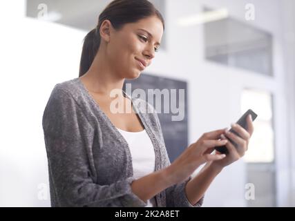 In jeder Minute des Tages in Kontakt bleiben. Eine schöne junge Frau, die ihr Smartphone im Büro benutzt. Stockfoto