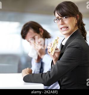 Eeuw. Eine Geschäftsfrau versucht, ihr Sandwich zu essen, während ein Mitarbeiter seine Nase bläst. Stockfoto