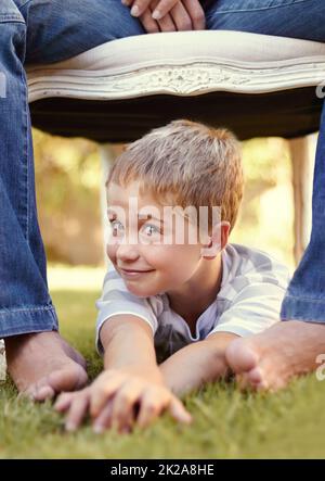 Ich denke, ich sollte seine Füße kitzeln. Aufnahme eines niedlichen kleinen Jungen, der auf dem Gras unter einem Stuhl liegt, auf dem sein Vater sitzt. Stockfoto
