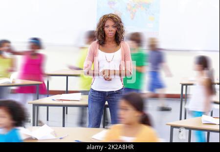 Scheut die Ruhe inmitten des Sturms. Porträt einer jungen Lehrerin mit ihren Schülern, die um sie herumlaufen. Stockfoto