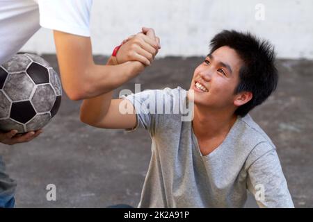 Eine helfende Hand geben. Asiatischer Junge, der seinem Freund während eines Fußballspiels vom Boden aus hilft. Stockfoto