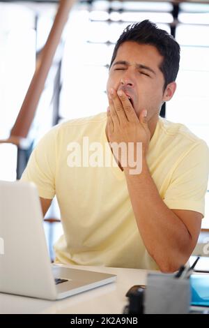 Zeit für meine nächste Tasse Kaffee. Ein junger Mann gähnt, während er an einem Schreibtisch vor einem Computer sitzt. Stockfoto