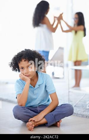 Das Gefühl, weggelassen zu werden Porträt eines traurig aussehenden kleinen Jungen, der zusammen mit seinen Schwestern im Hintergrund spielt. Stockfoto