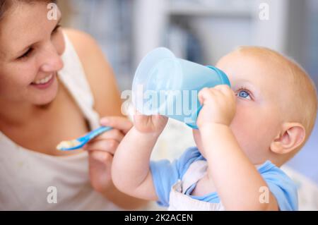 Trinkt ganz allein. Eine junge Mutter beobachtet, wie ihr kleiner Junge aus seinem sippy Cup trinkt. Stockfoto