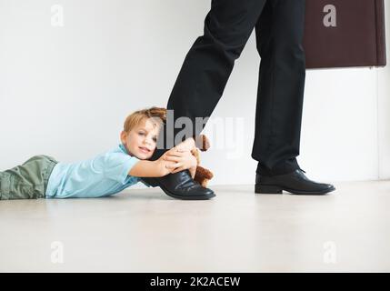 Trennungsangst in Aktion. Porträt eines niedlichen Jungen, der sich am Bein seines Dads hält, als er versucht, zur Arbeit zu gehen. Stockfoto