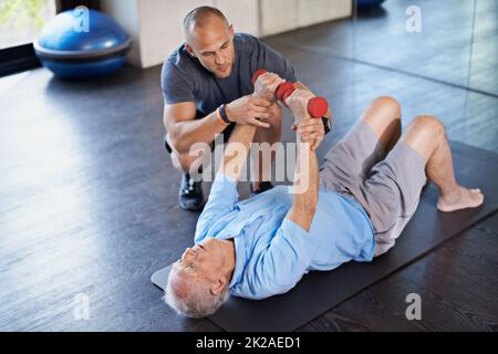 Er führt ihn zurück zum Wohlbefinden. Ein Physiotherapeut hilft einem älteren Mann mit Gewichten. Stockfoto
