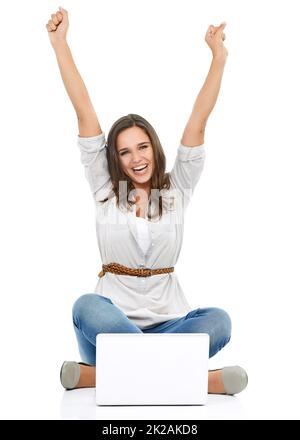 Tausend Treffer. Studioporträt einer jungen Frau, die vor einem Laptop sitzt und ihre Arme zur Feier angehoben hat. Stockfoto