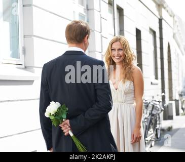 Hes eine solche neckerei. Eine schöne Frau lächelt ihren Freund an, der einen Haufen weißer Rosen hinter seinem Rücken hält. Stockfoto