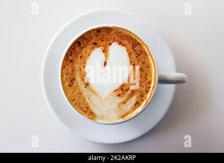 Der beste Start in jeden Morgen. Ein frisch zubereiteter Cappuccino auf einem weißen Tisch mit herzförmiger Form im Milchschaum. Stockfoto