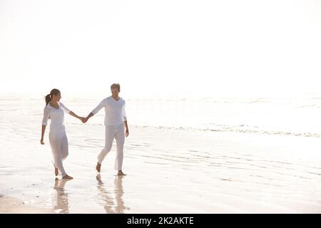 Hände durch die Schönheit der Natur halten. Ganzkörperaufnahme eines attraktiven jungen, weiß gekleideten Paares, das an einem Strand entlang läuft. Stockfoto