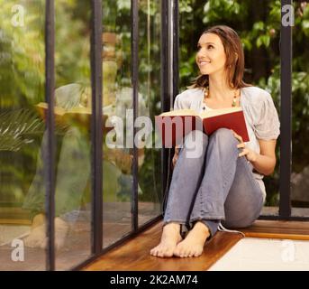 Dies ist mein Lieblingsleseport. Eine schöne Frau, die neben einem Fenster sitzt und liest. Stockfoto