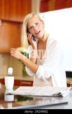 Die Hektik des modernen Lebens. Eine reizende junge Frau, die einen Anruf entgegennahm, während sie Tee machte und die Zeitung las. Stockfoto