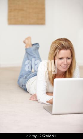 Kontakt mit Online-Freunden aufnehmen. Eine schöne reife Frau, die auf ihrem Laptop arbeitet, während sie auf dem Boden liegt. Stockfoto