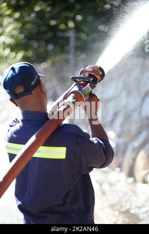 Feuerwehrmann bei der Arbeit. Rückansicht eines Feuerwehrmans, der Wasser mit einem Löschschlauch spritzt. Stockfoto