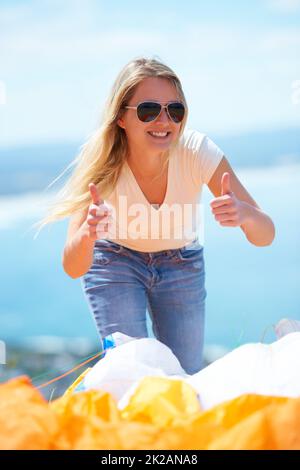 Sprungerfolg. Eine schöne junge Frau, die im Freien zwei Daumen nach oben gibt, mit einem Fallschirm im Vordergrund. Stockfoto