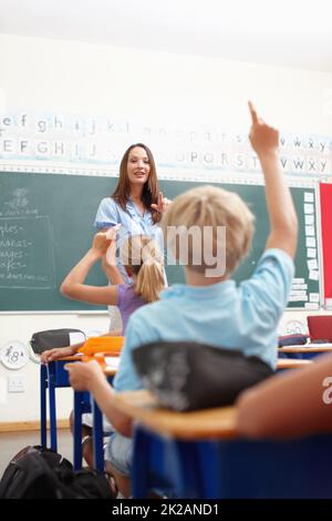 Ich habe eine Frage.... Eine hübsche junge Lehrerin, die an der Tafel der Klasse steht und unterrichtet. Stockfoto