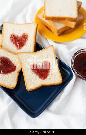 Toast, auf dem das Herz aus Marmelade besteht. Überraschendes Frühstückskonzept im Bett. Romantik für St. Valentinstag. Stockfoto