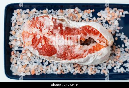 Frisches rohes Lachssteak mit grobem rosafarbenem Salz, zubereitet zum Backen auf dem Grill, liegt auf einem blauen Teller. Gesunde Meeresfrüchte. Draufsicht, Platz für eine Inschrift. Stockfoto